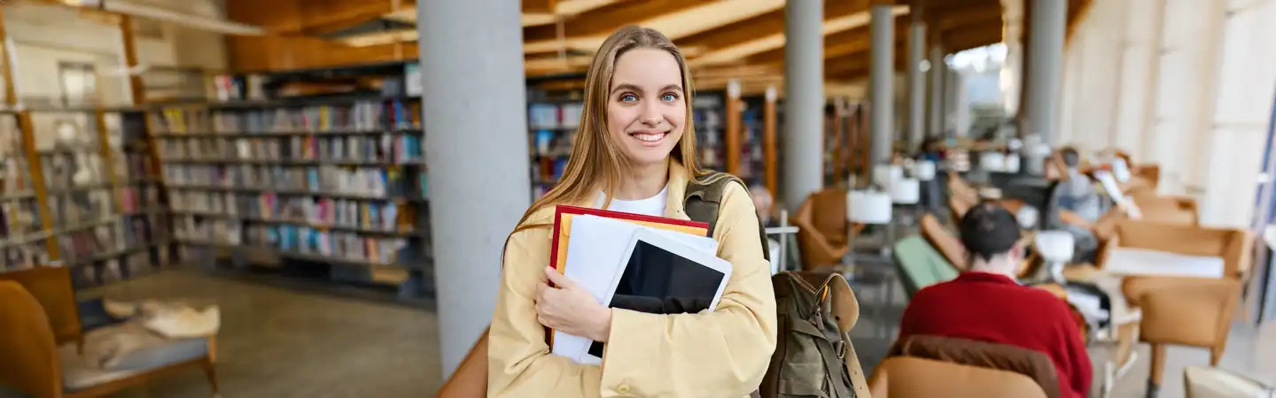 Smiling young woman holding books and a laptop in a library with shelves and people reading in the background.