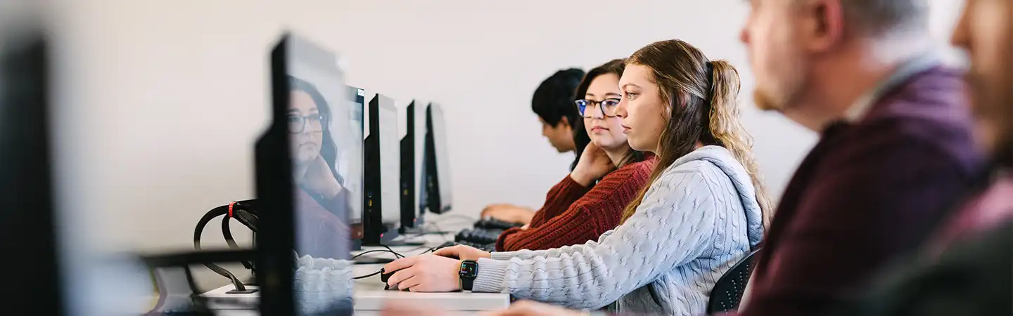 Four people working on desktop computers in a row, focused on their screens in a classroom or office setting.
