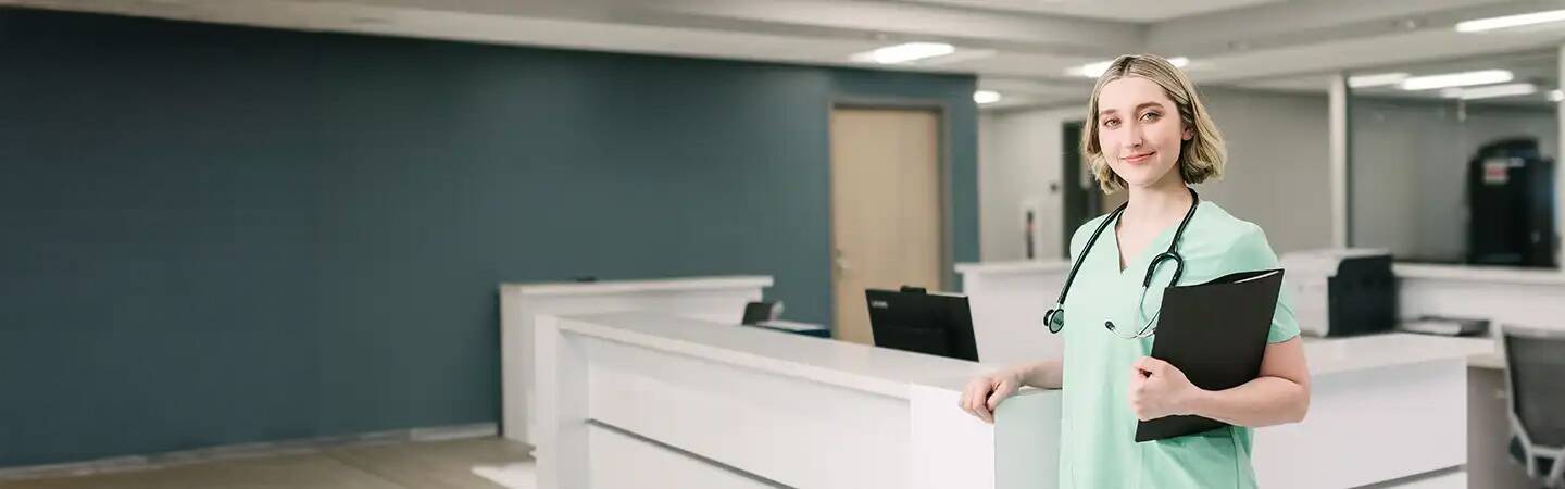 A nurse in scrubs stands with a clipboard in a modern, brightly lit hospital reception area.