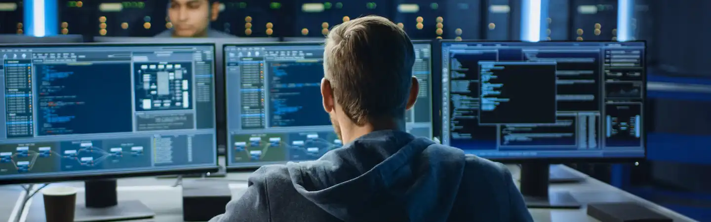 A person sits in front of multiple computer monitors displaying code and data in a dimly lit room.