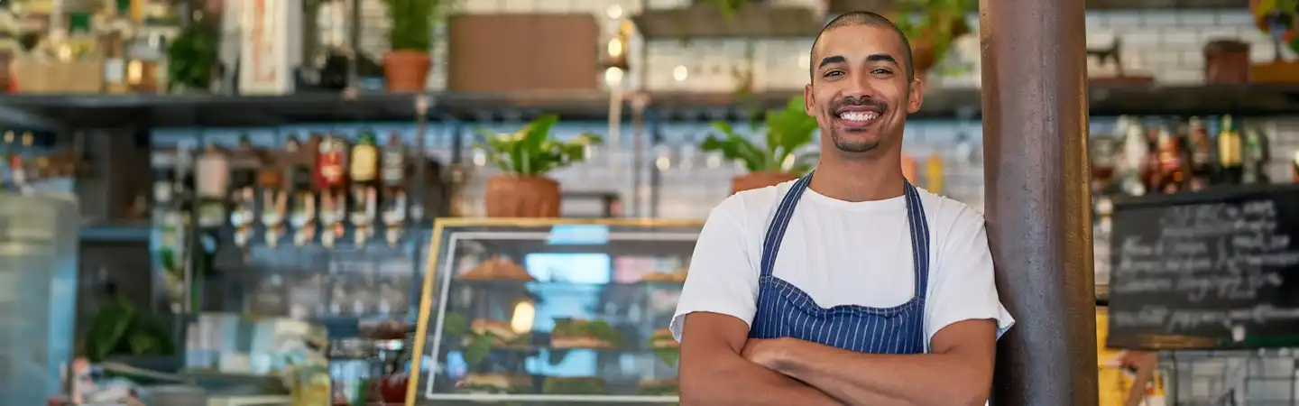 Smiling man in apron stands with arms crossed in front of a counter in a cozy cafe with plants and pastries.