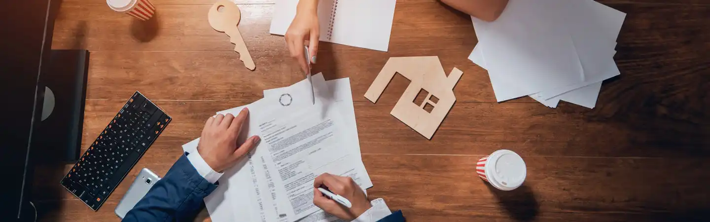 Two people signing a contract at a wooden desk with a house cutout, key cutout, papers, keyboard, and coffee cups.