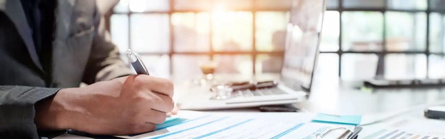 A person in a suit writes on a document at a desk with a laptop and other office supplies, with sunlight in the background.