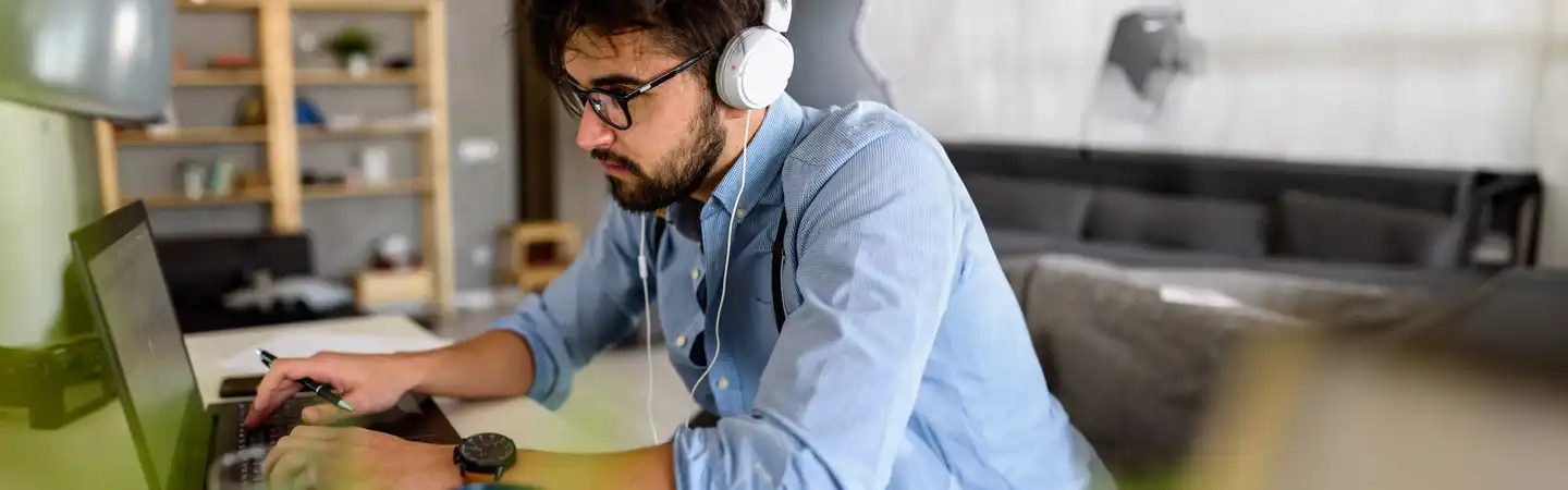 Man with glasses and headphones working on a laptop at a desk in a casual home office setting.