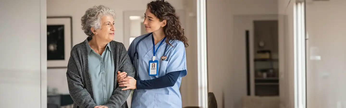A nurse in scrubs supports an elderly woman while they walk together indoors, sharing a warm and caring moment.