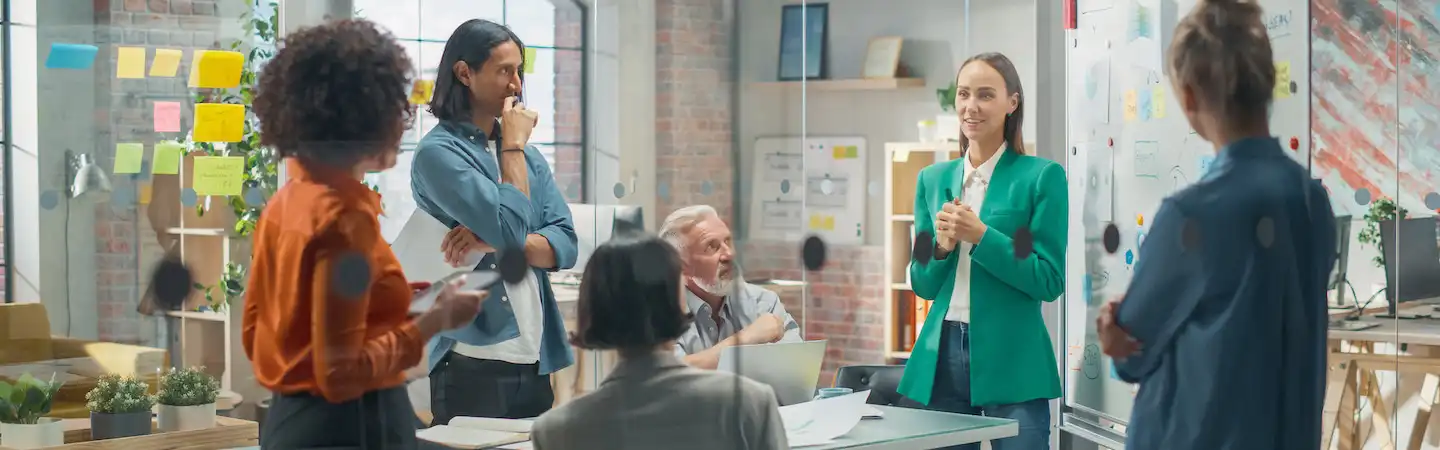 A diverse group of professionals engaged in a meeting in a modern office space with glass walls and sticky notes.