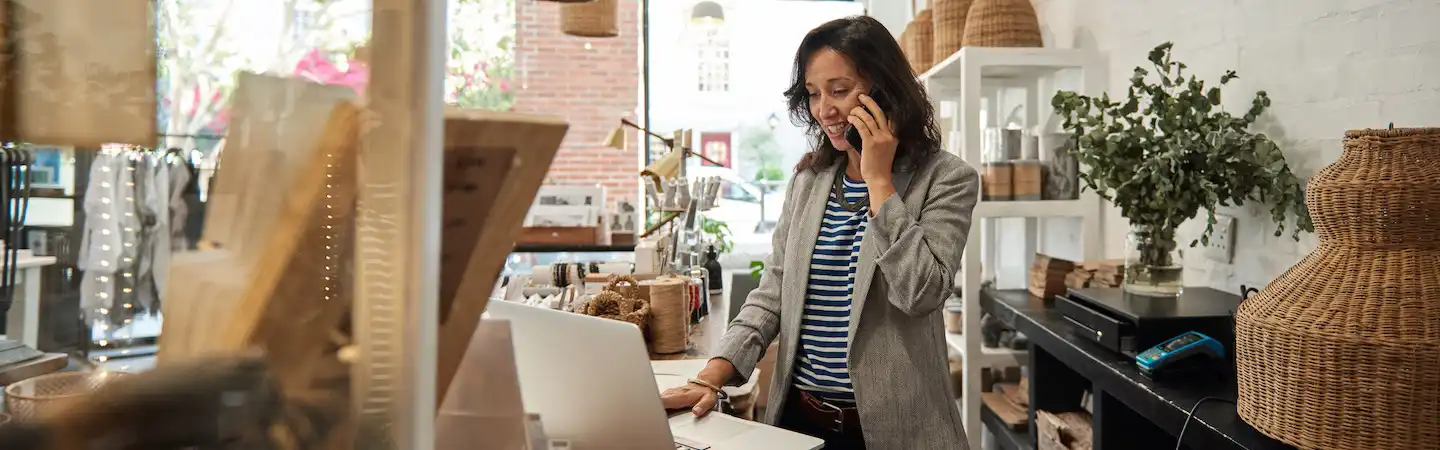 Woman in a store, talking on the phone while using a laptop, surrounded by various decor items and plants.