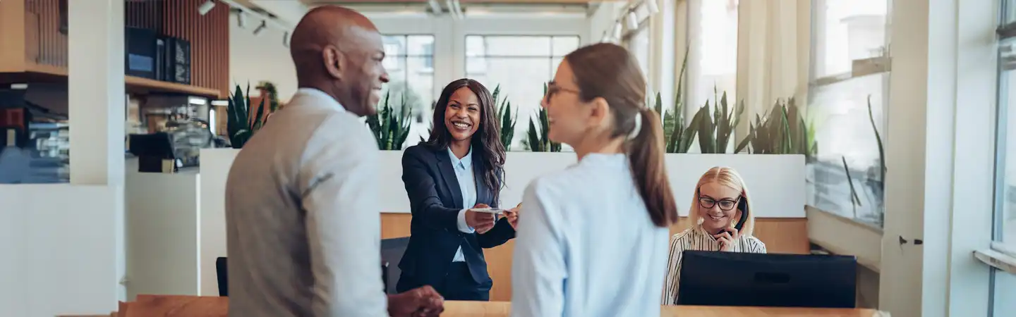 Four people in a bright office, two shaking hands, one smiling and holding a document, another working at a desk.