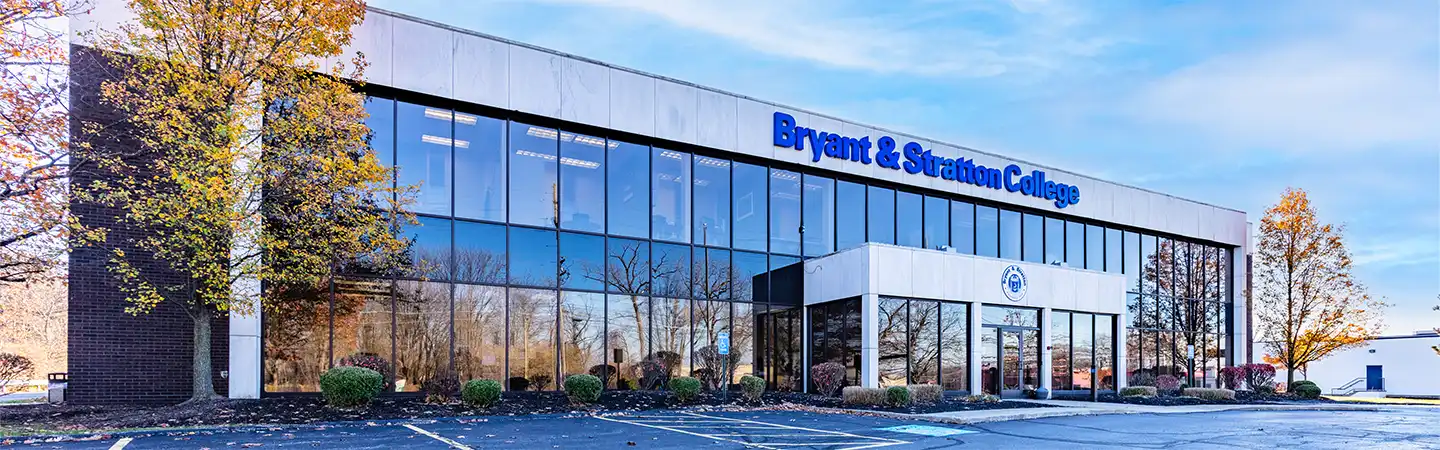 Exterior of Bryant & Stratton College building with large glass windows, surrounded by shrubs and trees with autumn leaves.