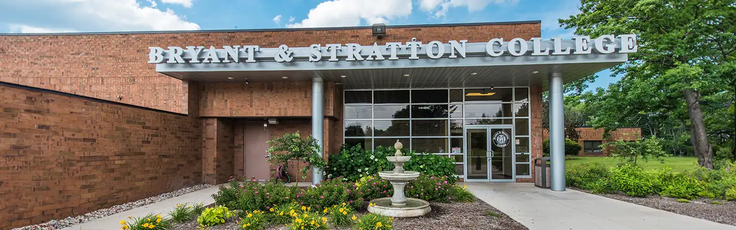 Entrance to Bryant & Stratton College building with brick facade, glass doors, and a small garden with a fountain.