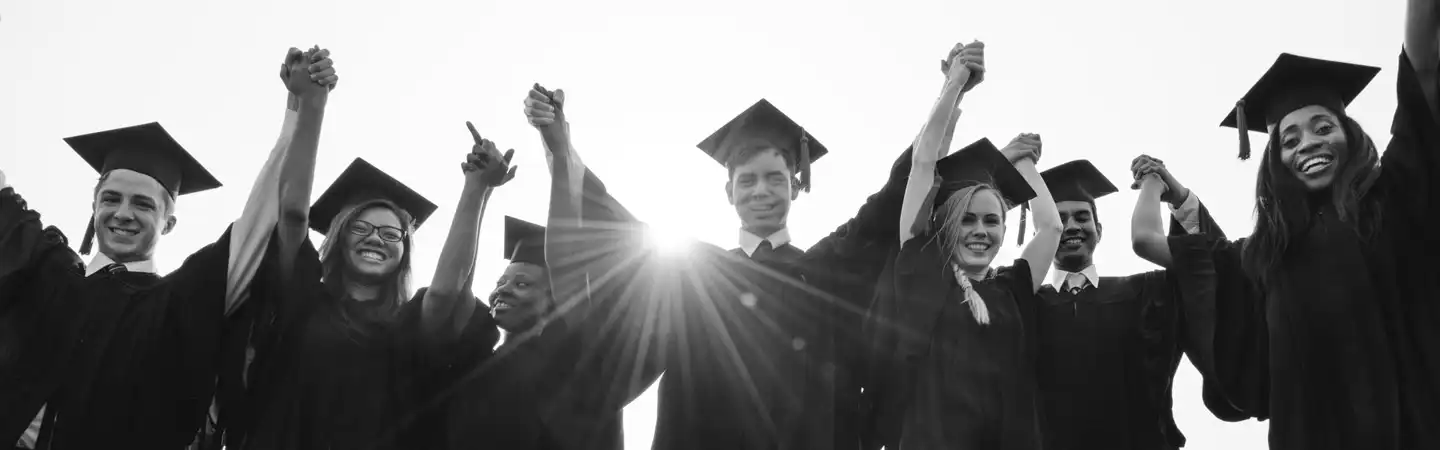 Grads in caps and gowns, holding hands and celebrating, with the sun shining behind them. Black and white photo.