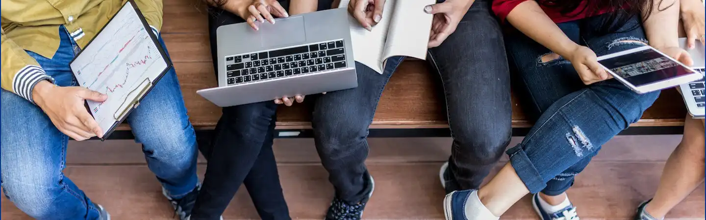 Four people seated, working on laptops, notebook, tablet, and clipboard with graphs, capturing a collaborative work setting.