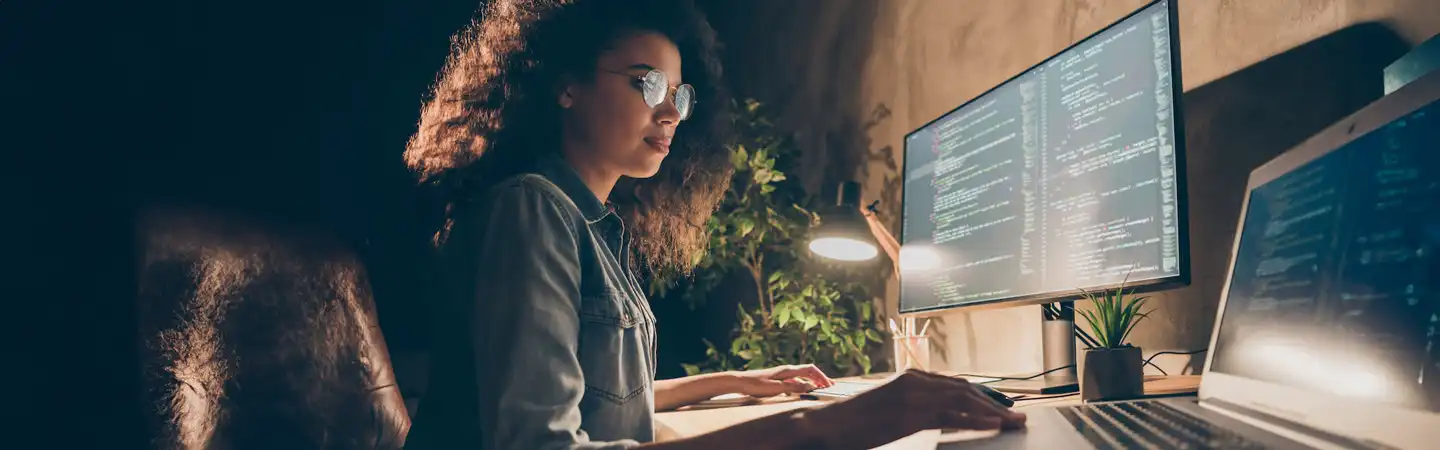 Person working at a desk with multiple screens displaying code, in a dimly lit room with a small plant nearby.