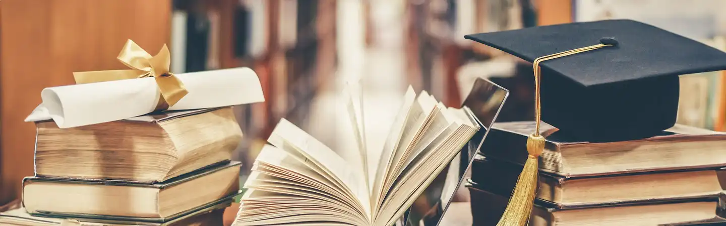Open books, rolled diploma tied with a ribbon, and a graduation cap on a stack of books in a library setting.