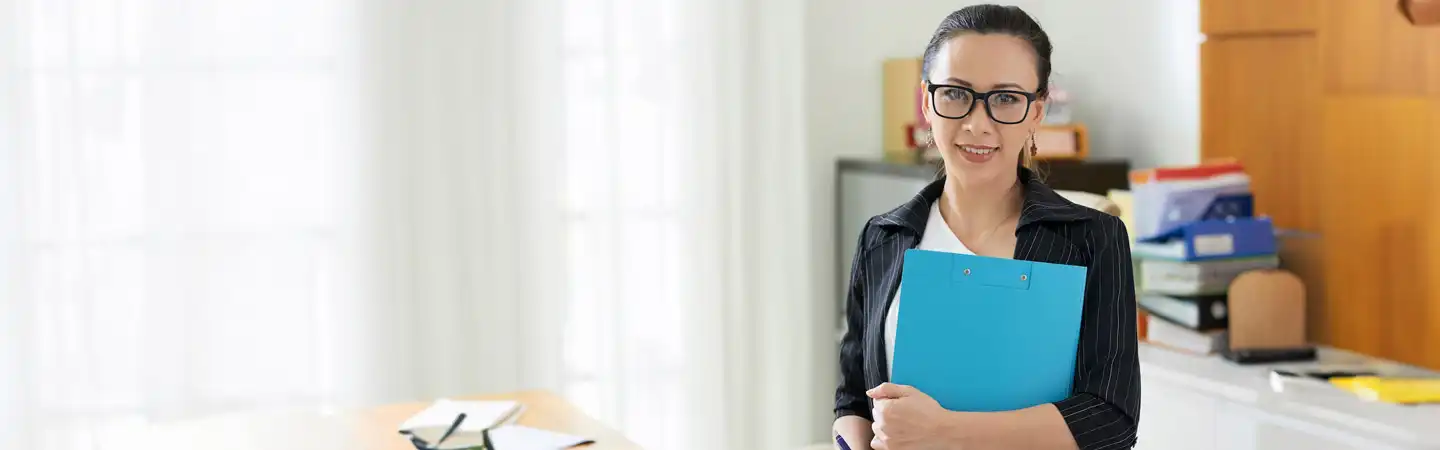 Smiling person in an office holding a blue folder, wearing glasses and a black jacket over a white top.