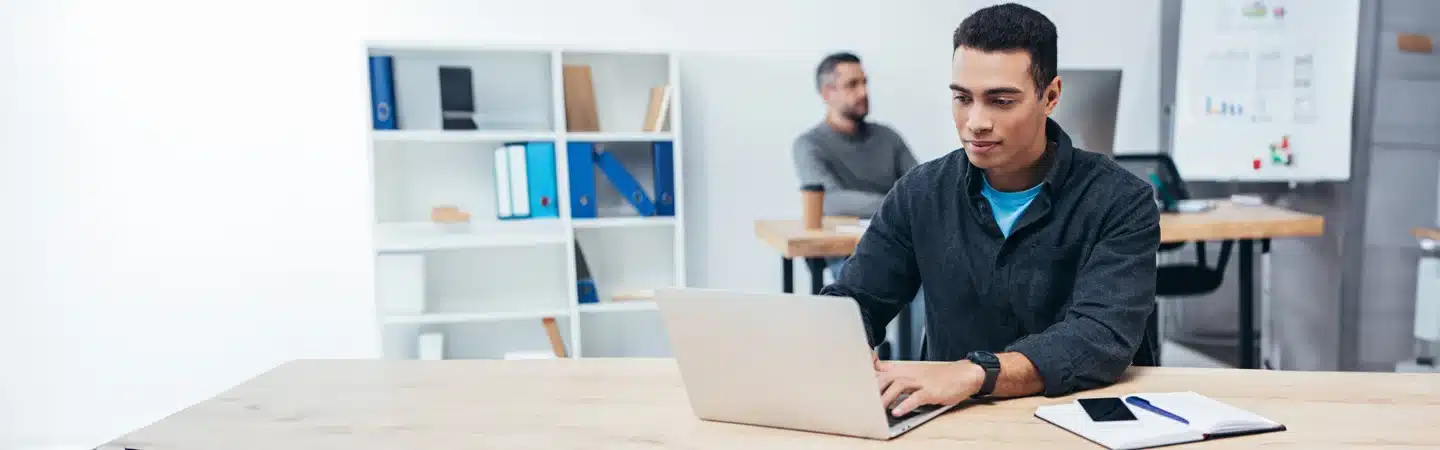 Man typing on a laptop at a shared office desk; another person works at a desk in the background with shelves nearby.