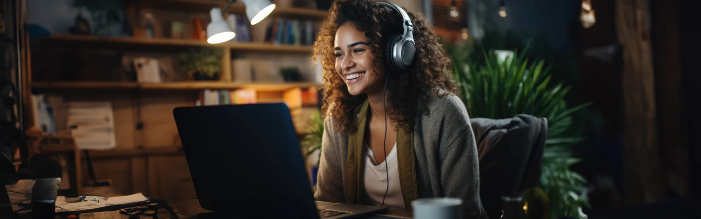 A person wearing headphones smiles while using a laptop in a cozy room with bookshelves and plants.