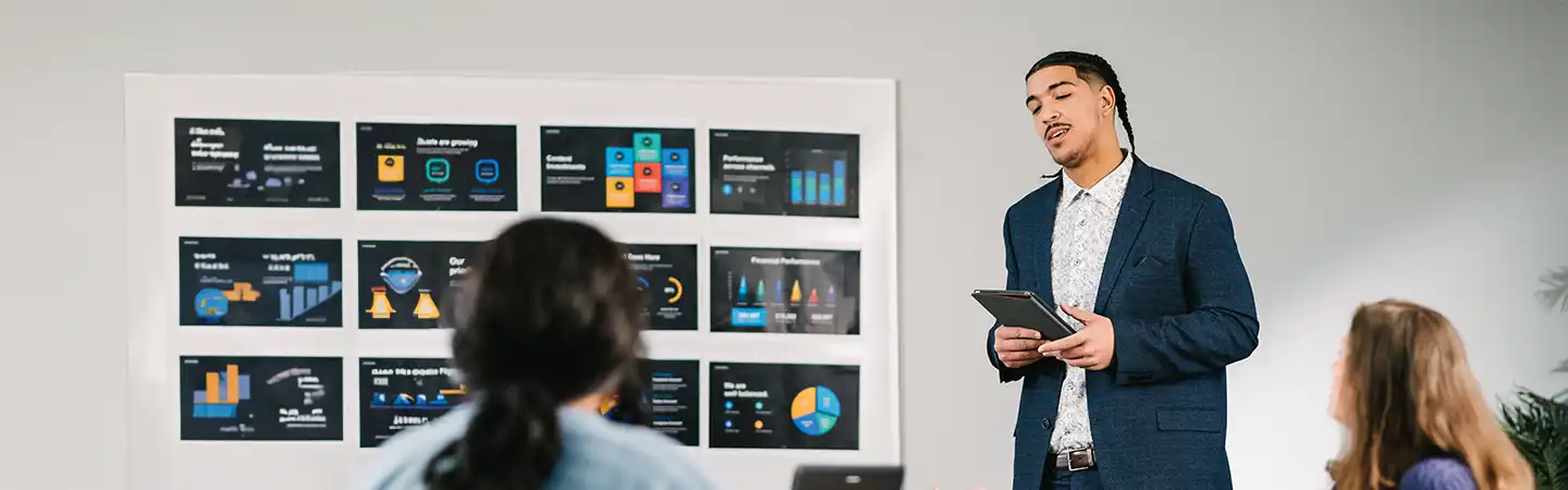 A person in a suit presents data from charts on a whiteboard to colleagues during a meeting in a modern office.