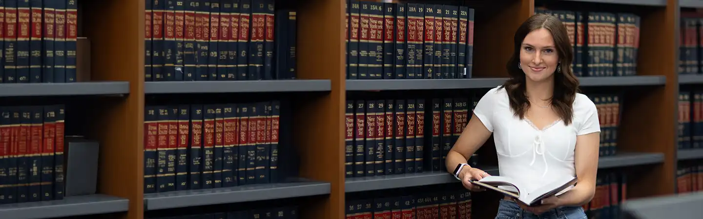 A woman reads a book while standing in front of shelves filled with law books in a library.