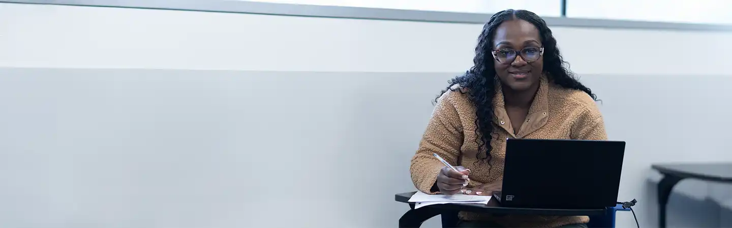 Person smiling while working on a laptop and writing in a notebook at a desk in a room with a large window.