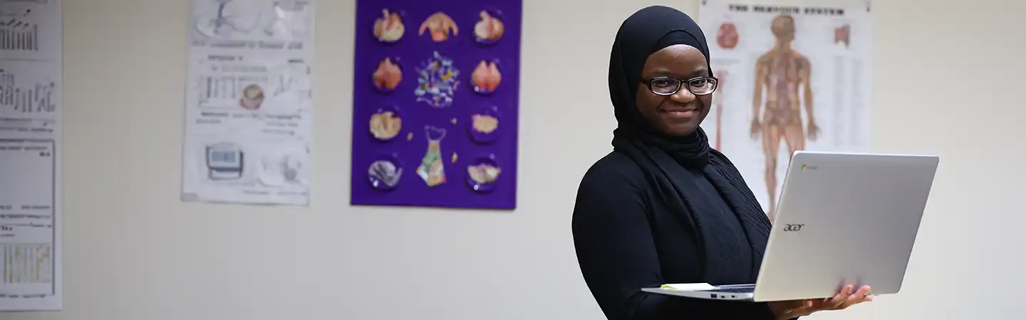 A person in black attire smiles while holding a laptop, with anatomy posters in the background.