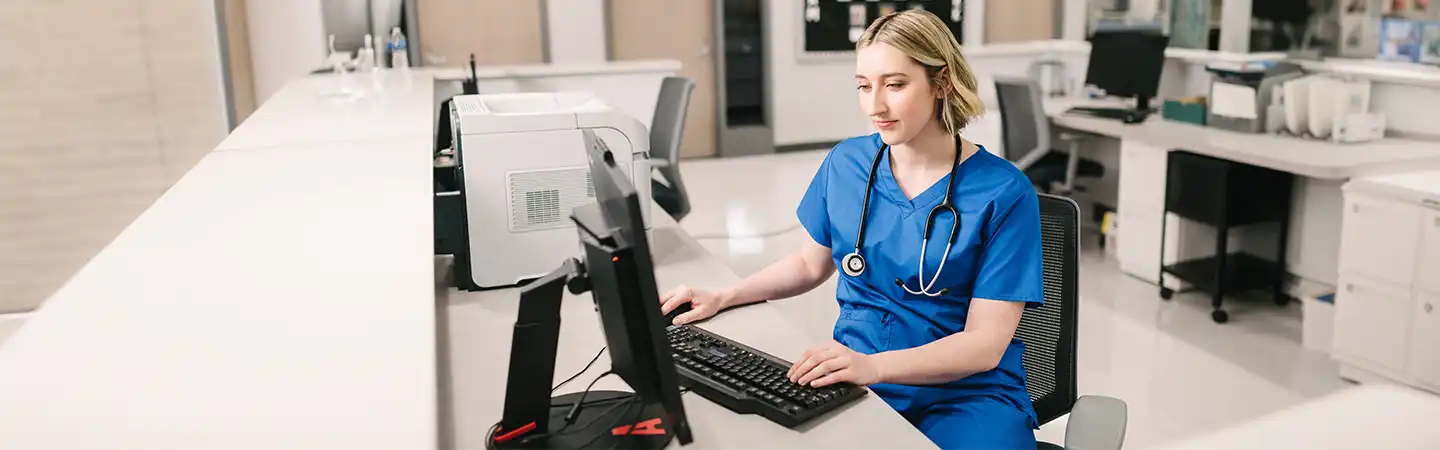 A healthcare worker in blue scrubs and a stethoscope works at a computer in a medical office or hospital.