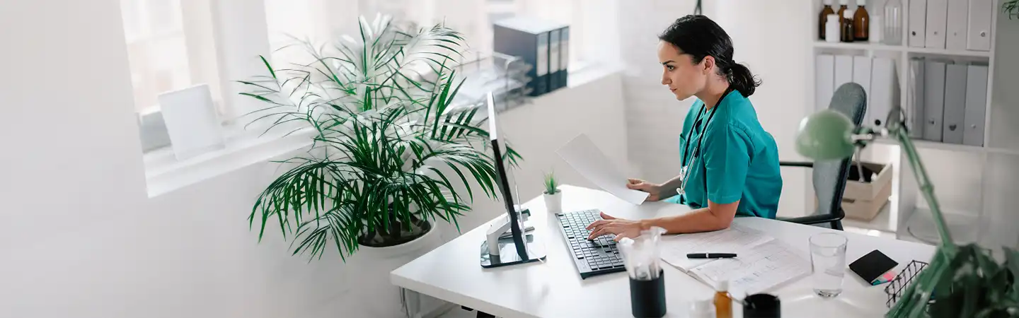 A woman wearing scrubs sits at a desk in a well-lit office, working on a computer, surrounded by plants and paperwork.