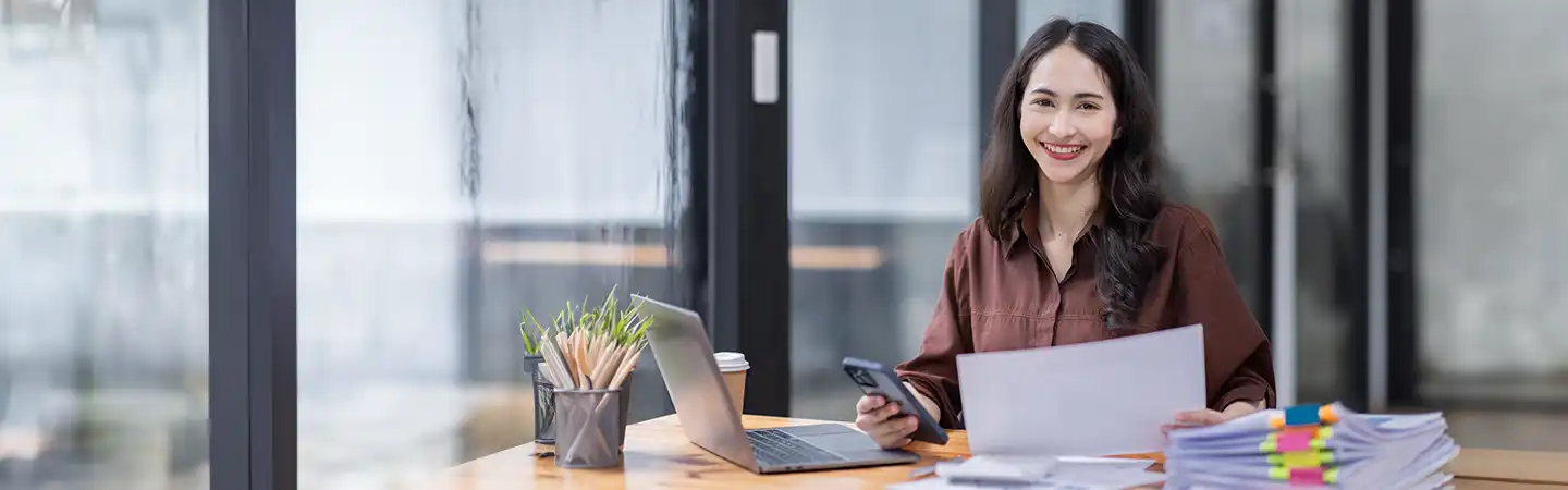 A woman smiling at her desk, holding a phone and papers, with a laptop and stacks of documents in front of her.