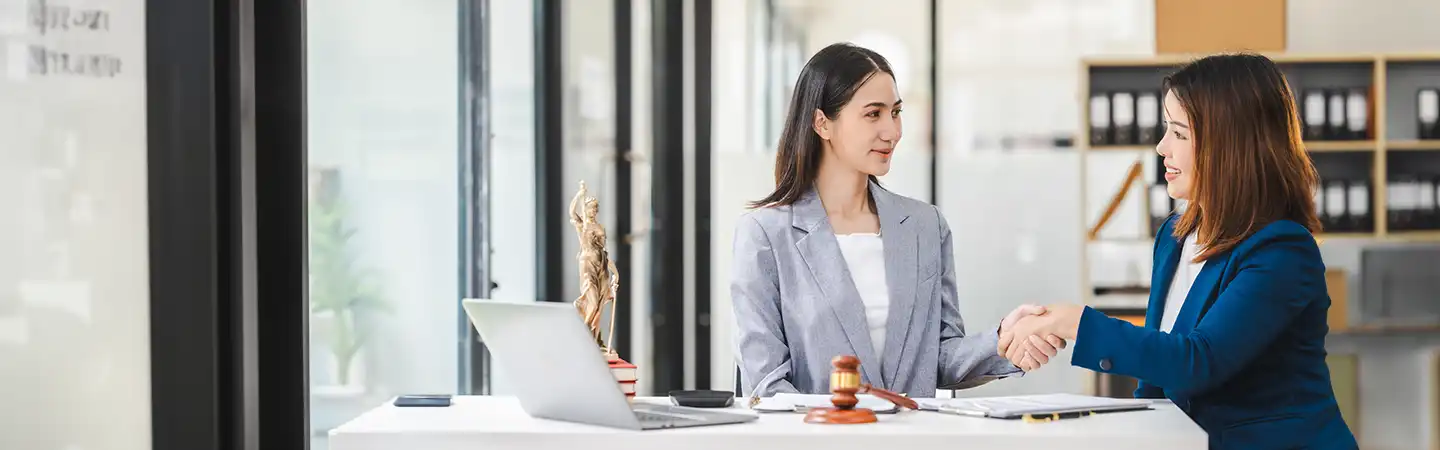Two women in business attire shaking hands across a desk with a laptop and gavel in an office setting.