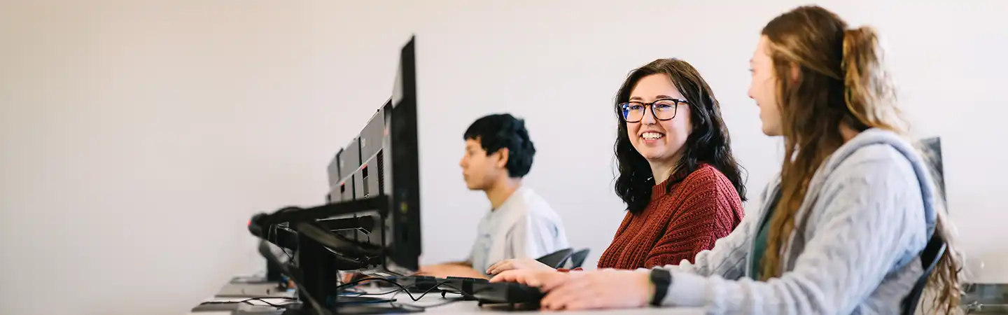 Three students work on computers in a classroom; two converse while the third is focused on his screen.