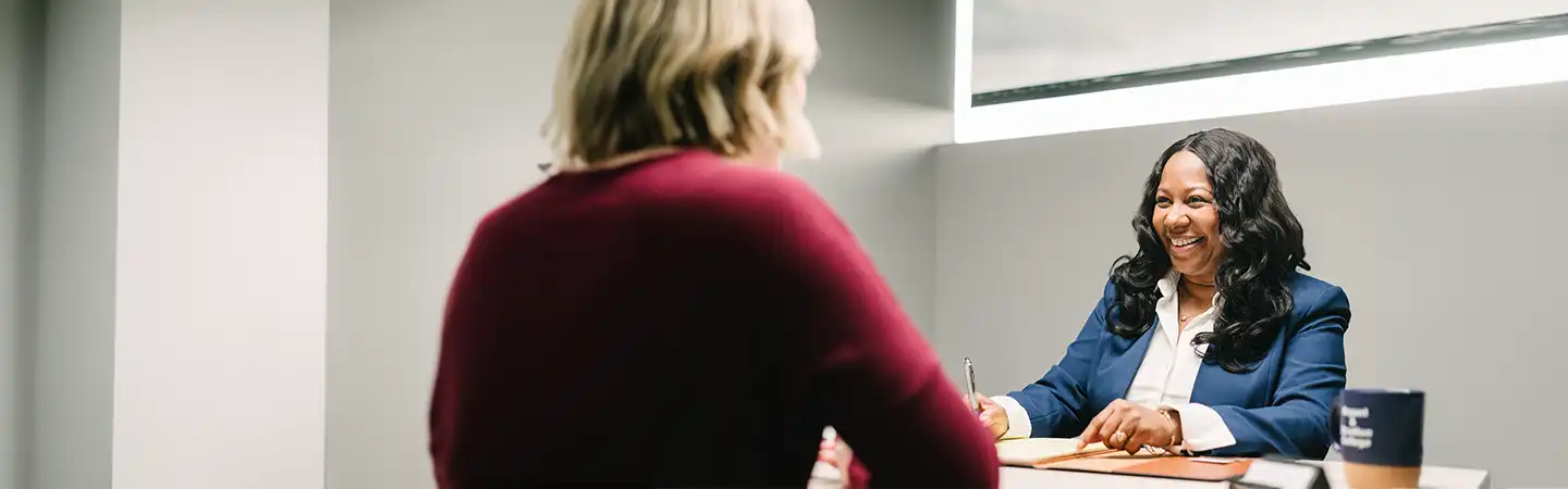 Two women in a professional setting; one, with dark hair in a blue blazer, smiles and takes notes opposite the other.