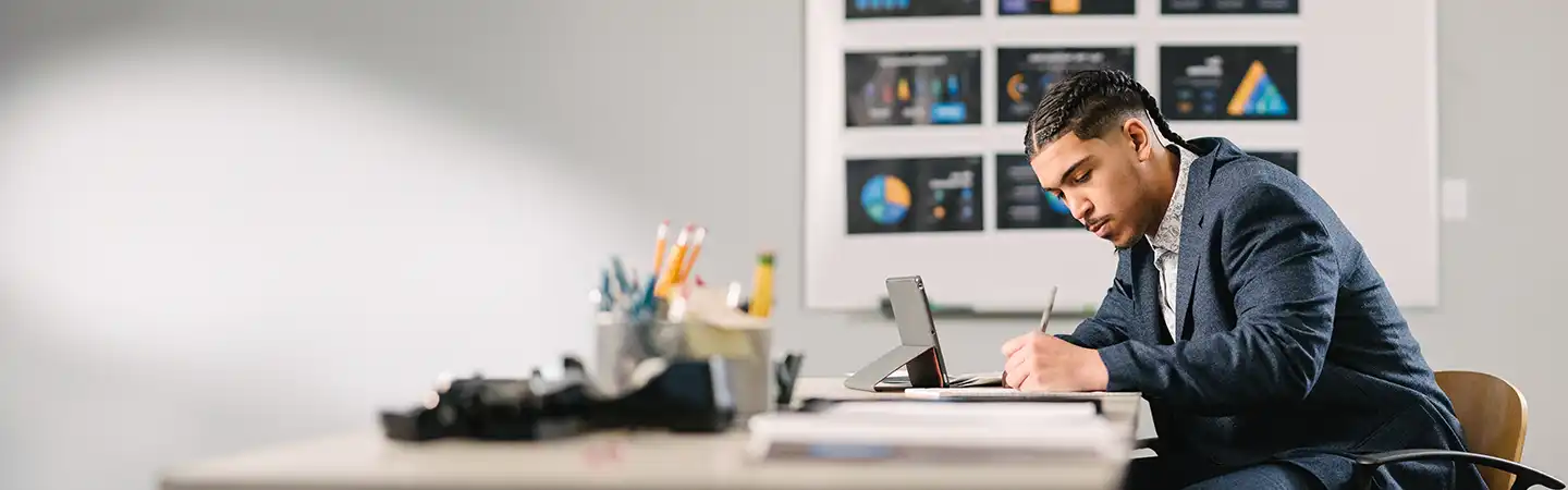 A man in a suit is sitting at a desk, writing on paper, with a laptop and charts on the wall in the background.