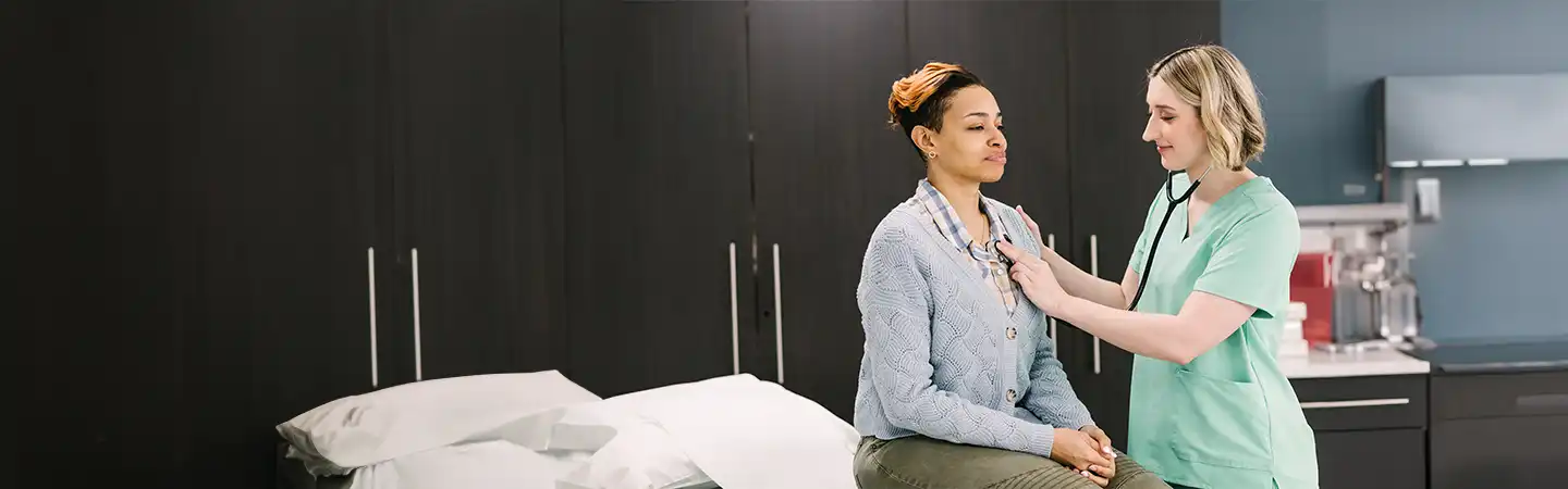 Healthcare professional in green scrubs examining a seated patient in a medical office.