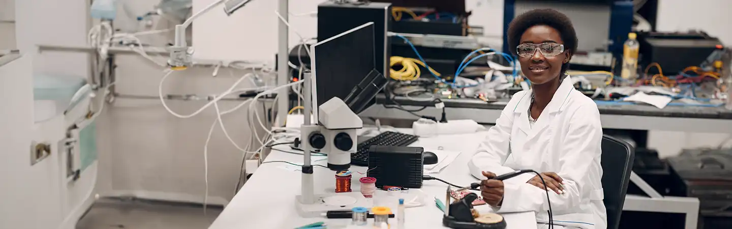 A person wearing a lab coat and safety glasses works with electronic equipment and a microscope in a laboratory setting.