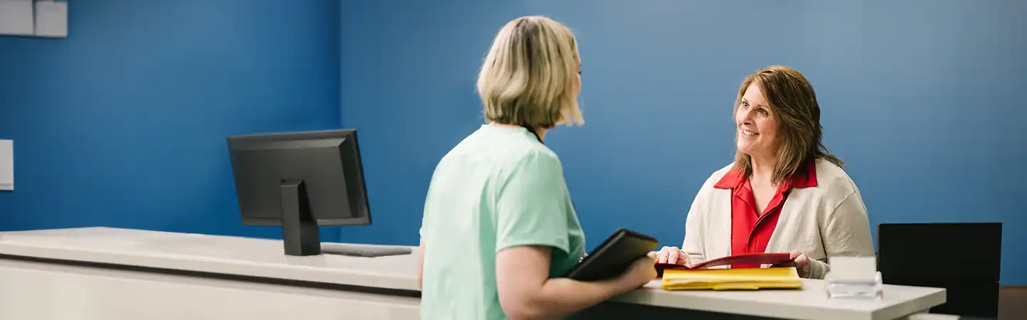 A woman in a green outfit speaks to another woman behind a reception desk with a computer monitor and paperwork.