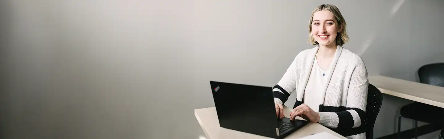 Person sitting at a desk and typing on a laptop with a smile, in a simple, well-lit room.
