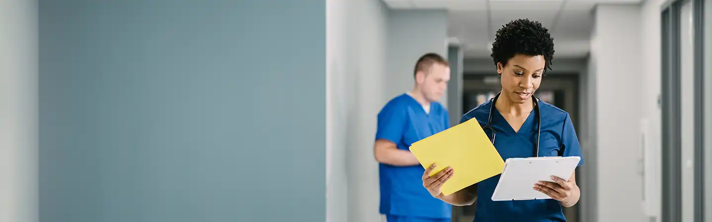 Two healthcare professionals in scrubs, one reading documents and another using a tablet, in a hospital hallway.