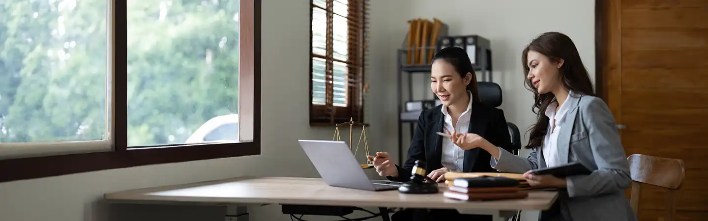 Two women in business attire discuss something on a laptop at a desk with legal books, a gavel, and scales.