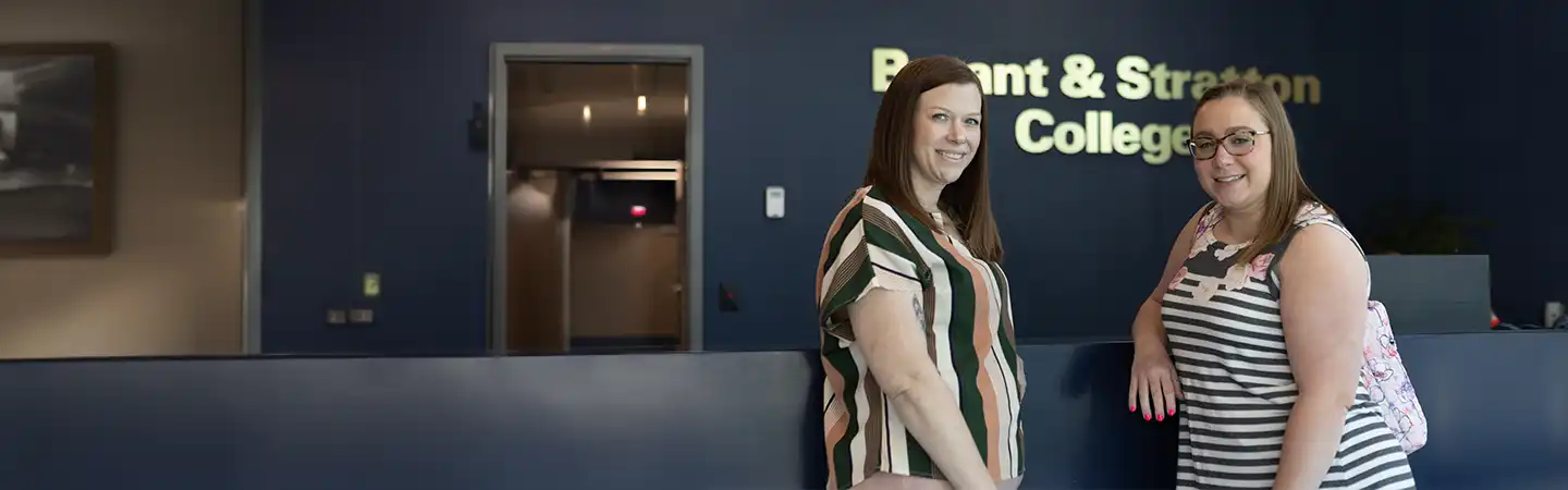 Two women stand smiling together in front of a reception desk with a "Bryant & Stratton College" sign on the wall behind them.