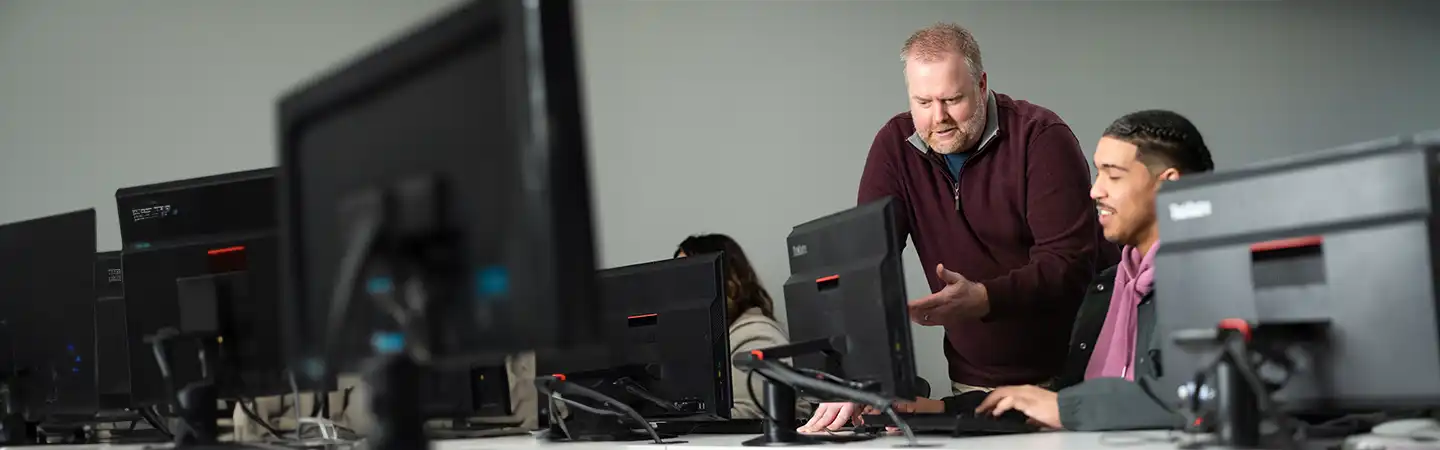 Instructor guides a student working on a computer in a classroom with multiple monitors.