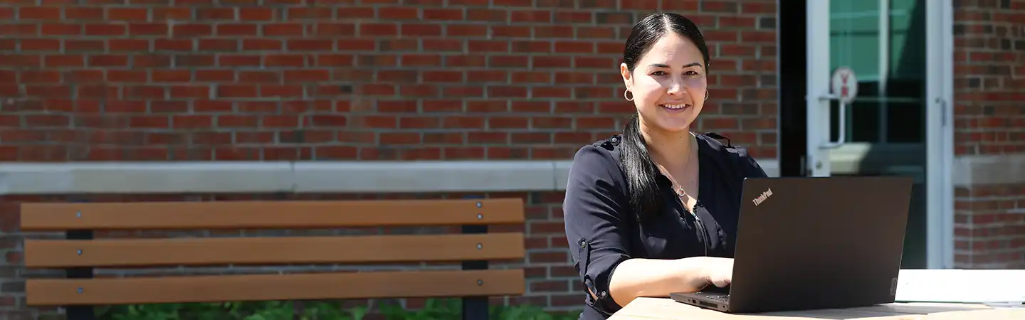 A person with long dark hair, wearing a black top, smiling while using a laptop outdoors on a wooden bench with a brick wall behind.