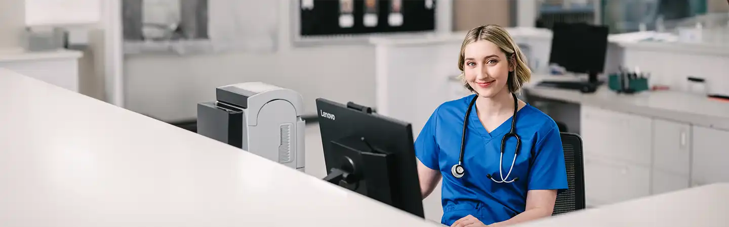 A nurse in blue scrubs sits at a desk with a computer, looking at the camera and smiling. A stethoscope hangs around her neck.