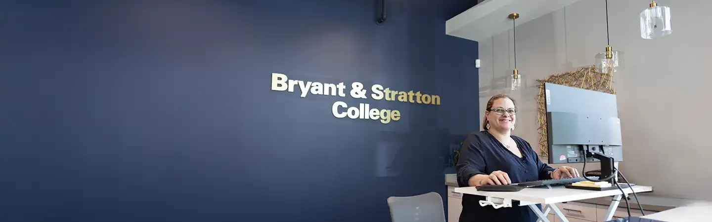 Person working at a desk with a computer in front of a wall with the sign "Bryant & Stratton College" on a dark blue background.
